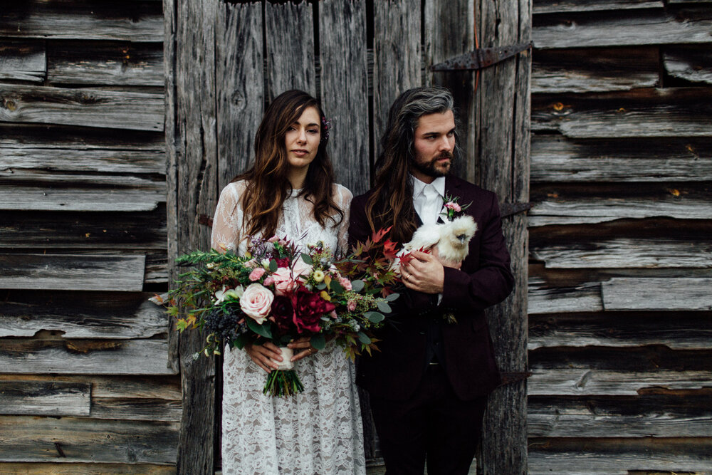 Bride in BHLDN wedding dress and groom with long hair wearing a maroon suit holding a chicken after their farmhouse wedding.