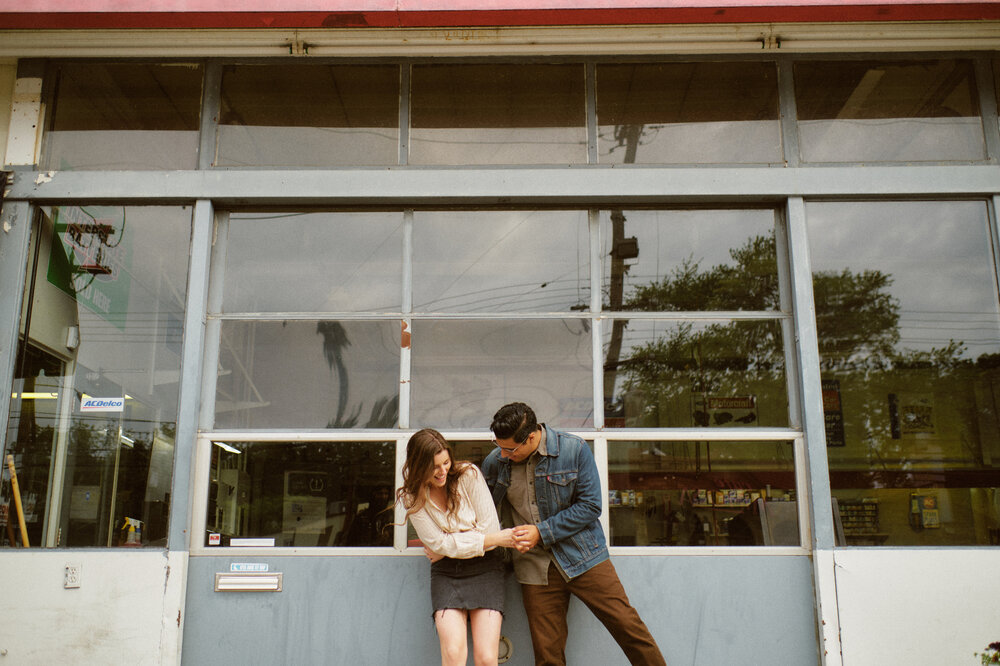 Photo of young couple in front of a mechanic garage holding hands and laughing on Bardstown Road in Louisville, Kentucky.