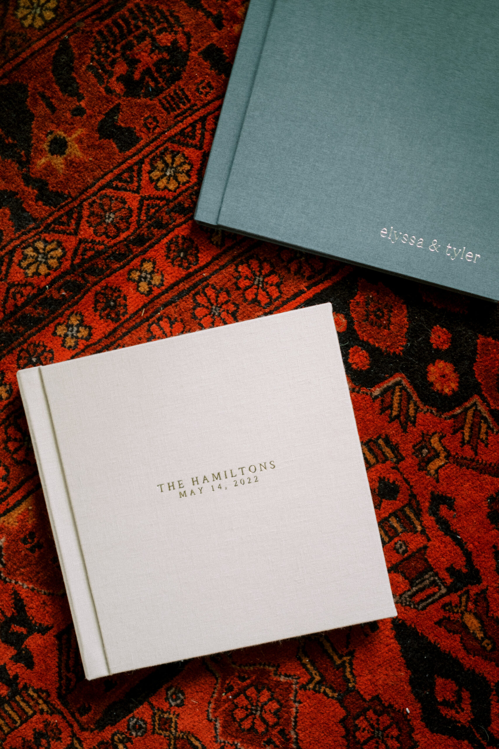 Two heirloom wedding albums, one a cream color with a gold serif title, and the other a dark dusty blue with silver serif text, lay on a red turkish rug.